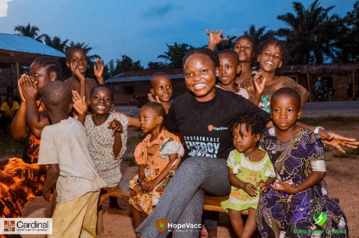 A Solfa Power team member in a branded shirt with a group of smiling children at dusk, lit by reliable clean energy